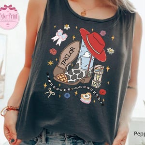 May include: A dark gray sleeveless tank top featuring a graphic print of a cow-print cowboy boot, a red hat, a beaded bracelet with the text "I KNEW YOU WERE", and blocks spelling "BTS". The boot includes the name "TAYLOR" and the number "13".