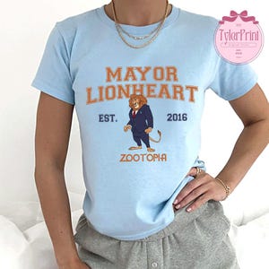 May include: Light blue t-shirt featuring the text "MAYOR LIONHEART" in orange, with an illustration of a lion in a suit. Below the text is "ZOOTOPIA" in orange, with "EST. 2016" in blue. The shirt is a crew neck.