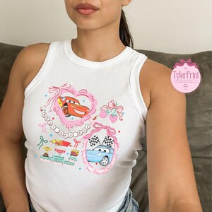 Puede incluir: Camiseta blanca de tirantes con un dise&ntilde;o gr&aacute;fico rosa y blanco con dos coches de dibujos animados. El gr&aacute;fico incluye el texto "Best Driver Ever" en un c&iacute;rculo rosa con una cinta rosa.