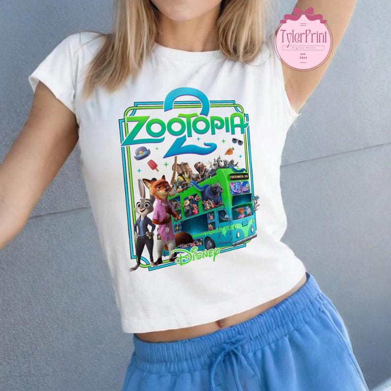 Puede incluir: Camiseta blanca con un gr&aacute;fico colorido de la pel&iacute;cula Zootopia 2. El dise&ntilde;o incluye el t&iacute;tulo de la pel&iacute;cula, personajes y un autob&uacute;s. El logotipo de Disney est&aacute; en la parte inferior del gr&aacute;fico.
