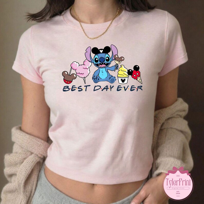 Pode incluir: Camiseta rosa com um personagem Stitch azul segurando um cone de sorvete do Mickey Mouse. A camiseta tem o texto "Best Day Ever" impresso.