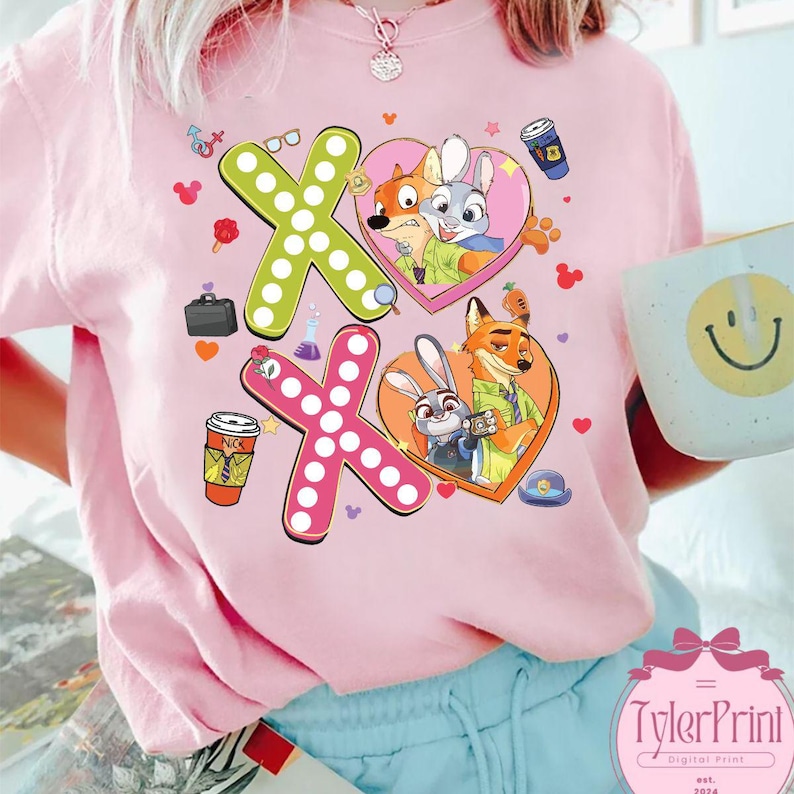 May include: Pink crew neck sweatshirt featuring a colorful graphic design with the characters Nick Wilde and Judy Hopps from Zootopia. The design includes the letters "XO" with heart and cartoon elements. The brand "TylerPrint" is visible at the bottom.