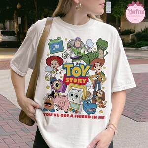 May include: White t-shirt featuring a colorful graphic of Toy Story characters, including Woody, Buzz Lightyear, and Rex. The words "TOY STORY" are prominently displayed, with the phrase "YOU'VE GOT A FRIEND IN ME" below. The shirt has a crew neck and short sleeves.