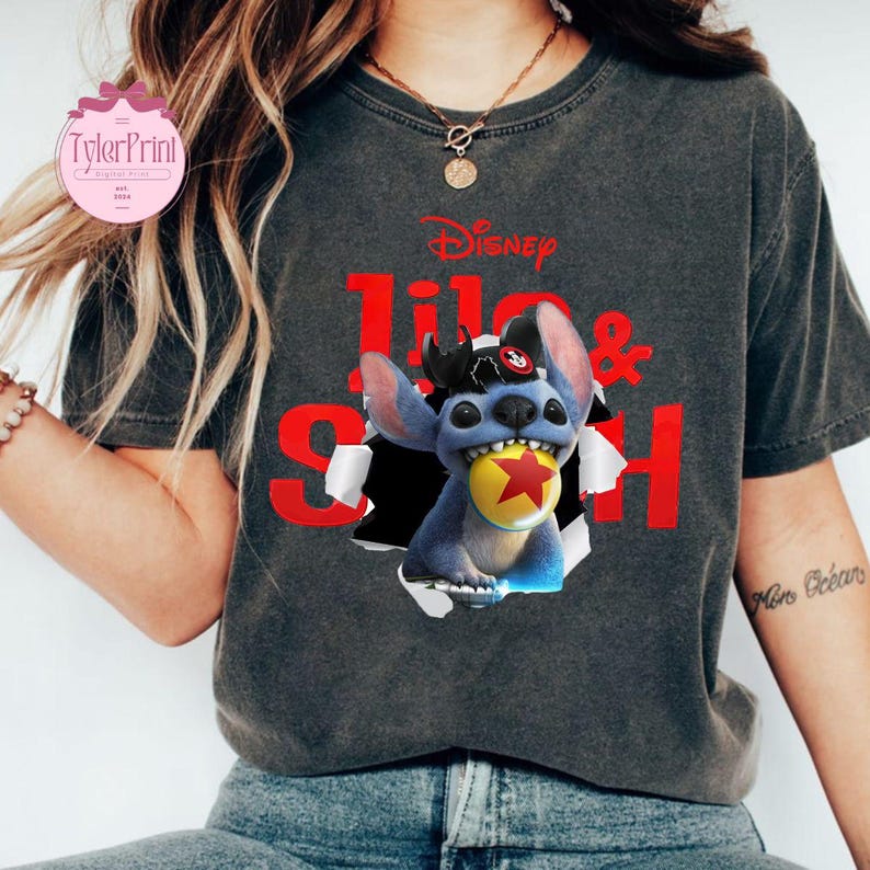 May include: Dark grey t-shirt with the Disney logo and the words "Lilo & Stitch" in red. The graphic depicts Stitch, a blue alien, breaking through a torn paper effect, holding a yellow ball with a red star.