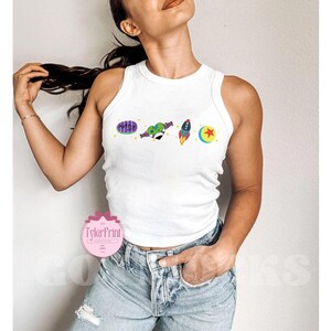 May include: White tank top with a graphic print of characters from the movie Toy Story. The graphic includes Woody, Buzz Lightyear, and the alien characters. The tank top is cropped and has a racerback style.