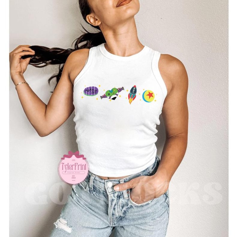 May include: White tank top with a graphic print of characters from the movie Toy Story. The graphic includes Woody, Buzz Lightyear, and the alien characters. The tank top is cropped and has a racerback style.