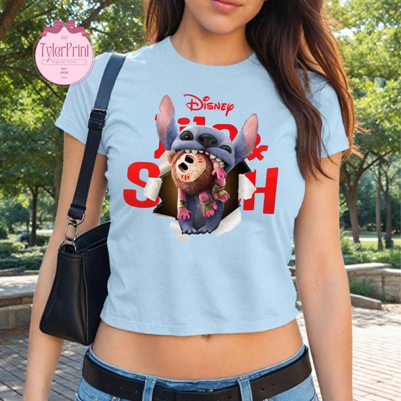 May include: Pale blue t-shirt with a graphic of Stitch from Disney's Lilo & Stitch. The design shows Stitch emerging from a torn hole, holding a brown stuffed animal. The word "Disney" and the word "Stitch" are in red.