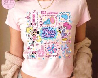 May include: A light pink t-shirt featuring a colorful graphic design with the words "UCA Nationals" and "Cheer Mom." The design includes cartoon characters, stars, a megaphone, and a bow. The shirt has a round neckline and short sleeves.