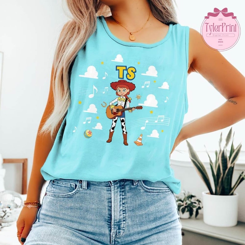 May include: A light blue sleeveless tank top featuring a graphic of a cartoon cowgirl playing an acoustic guitar. The design includes white clouds, musical notes, a yellow star, a ball, and an orange cone, with the letters TS printed above.