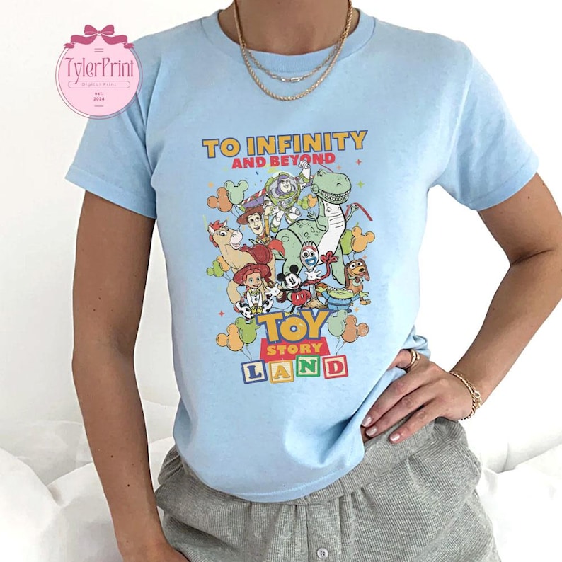 以下が含まれることがあります： 水色のTシャツ。カラフルなトイ・ストーリーのグラフィックがプリントされており、「TO INFINITY AND BEYOND」と「TOY STORY LAND」の文字が入っています。半袖で、クルーネックです。