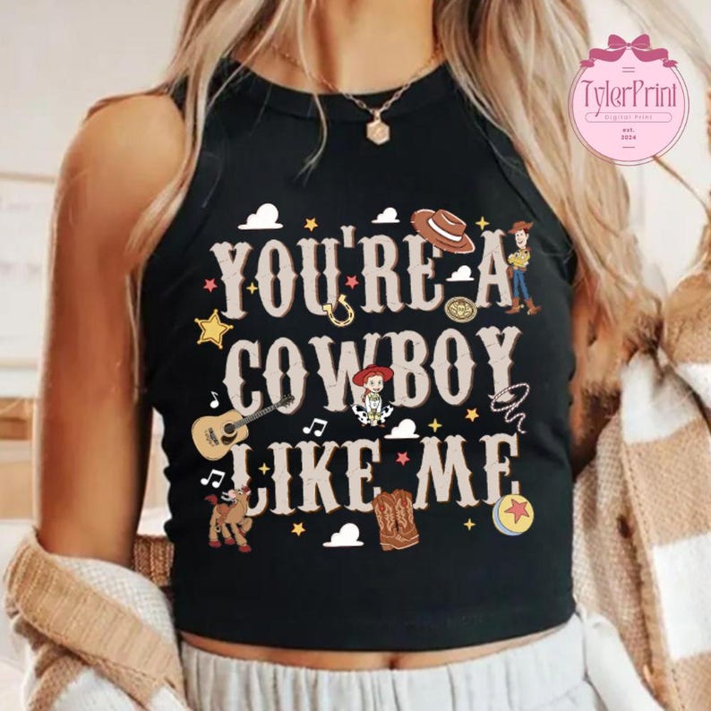 May include: A black sleeveless tank top featuring the text "YOU'RE A COWBOY LIKE ME" in a western-style font. The design includes illustrations of a cowboy hat, a guitar, a lasso, a star, a horse, boots, and a small cartoon character.