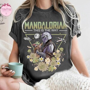 May include: Dark gray t-shirt featuring a graphic of The Mandalorian and Grogu surrounded by flowers and space-themed elements. The text "MANDALORIAN - THIS IS THE WAY" is printed across the top. The shirt has a vintage, worn look.