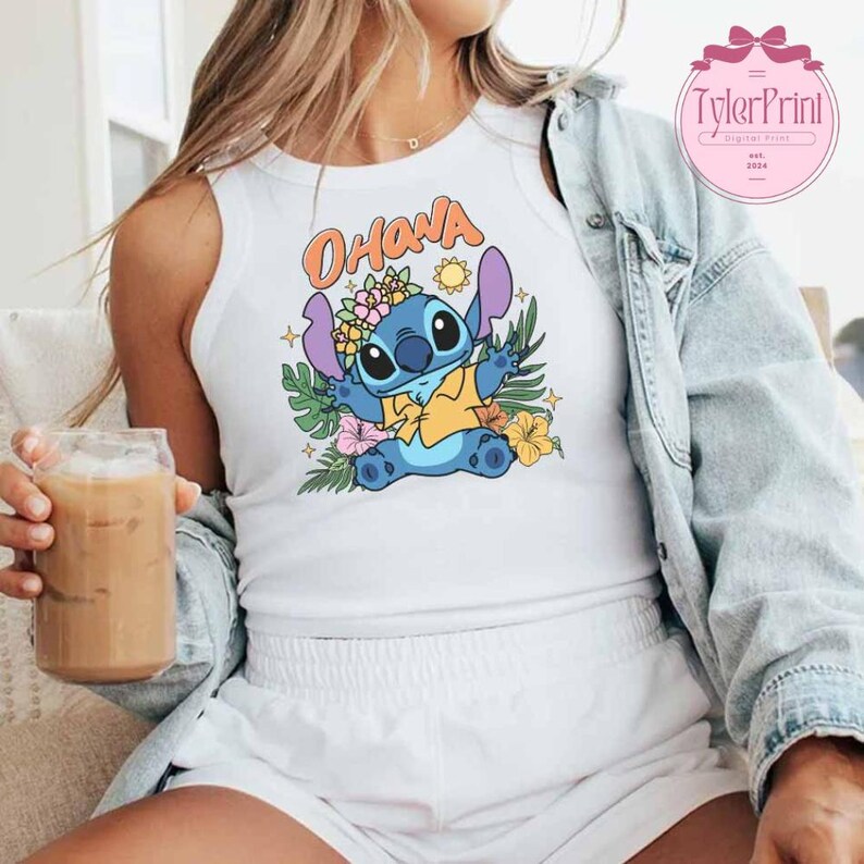 May include: White tank top featuring a cartoon Stitch character with the word "Ohana" in orange lettering. Stitch is surrounded by flowers and tropical leaves. The design is colorful and vibrant.