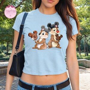 Puede incluir: Camiseta azul claro con una ilustraci&oacute;n de dibujos animados de dos ardillas listadas sosteniendo helados con forma de Mickey Mouse. Las ardillas listadas llevan sombreros y tienen la nariz roja. La camiseta tiene cuello redondo y mangas cortas.