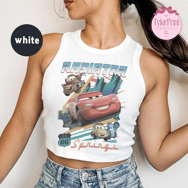 May include: A white sleeveless crop top featuring a graphic print of characters from the movie Cars. The design includes the text Radiator Springs and a US 66 road sign, with illustrations of Lightning McQueen, Mater, and other vehicle characters.