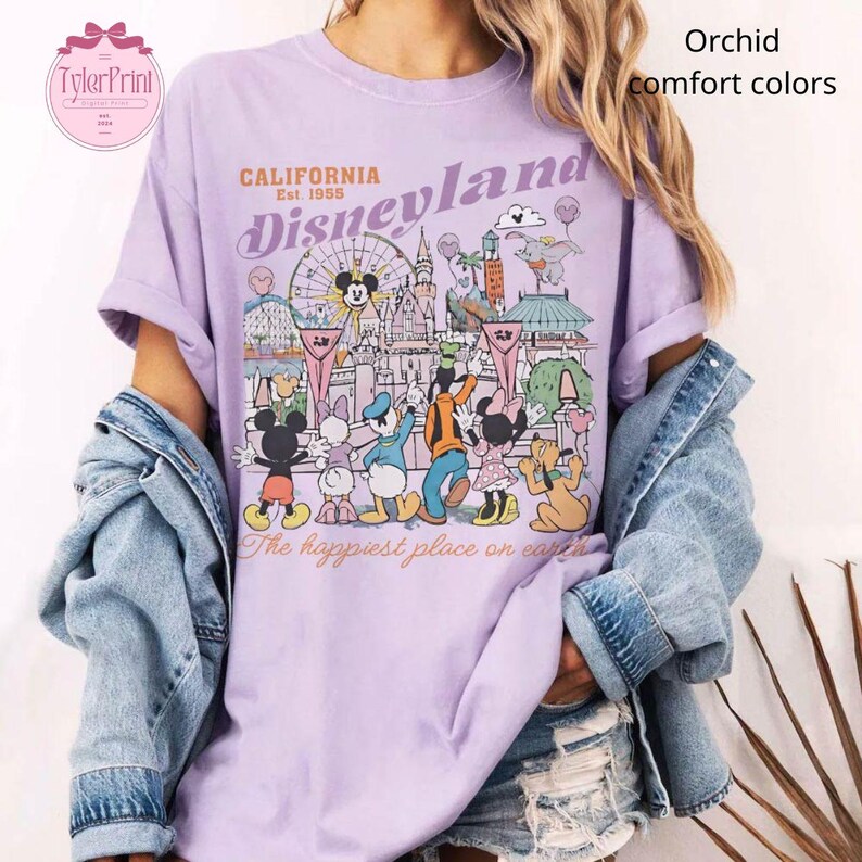 Puede incluir: Camiseta color orqu&iacute;dea con un gr&aacute;fico de Disneyland. El gr&aacute;fico presenta a Mickey Mouse, el Pato Donald, Goofy y Minnie Mouse frente a un castillo y una noria. El texto incluye "California Disneyland Est. 1955" y "The happiest place on earth."