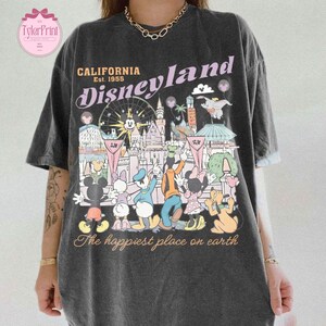 Puede incluir: Camiseta gris oscuro con un gr&aacute;fico colorido de Disneyland. El dise&ntilde;o incluye el texto "California Disneyland Est. 1955" y una representaci&oacute;n de varios personajes y lugares emblem&aacute;ticos de Disney. Tambi&eacute;n se incluye la frase "The happiest place on earth".