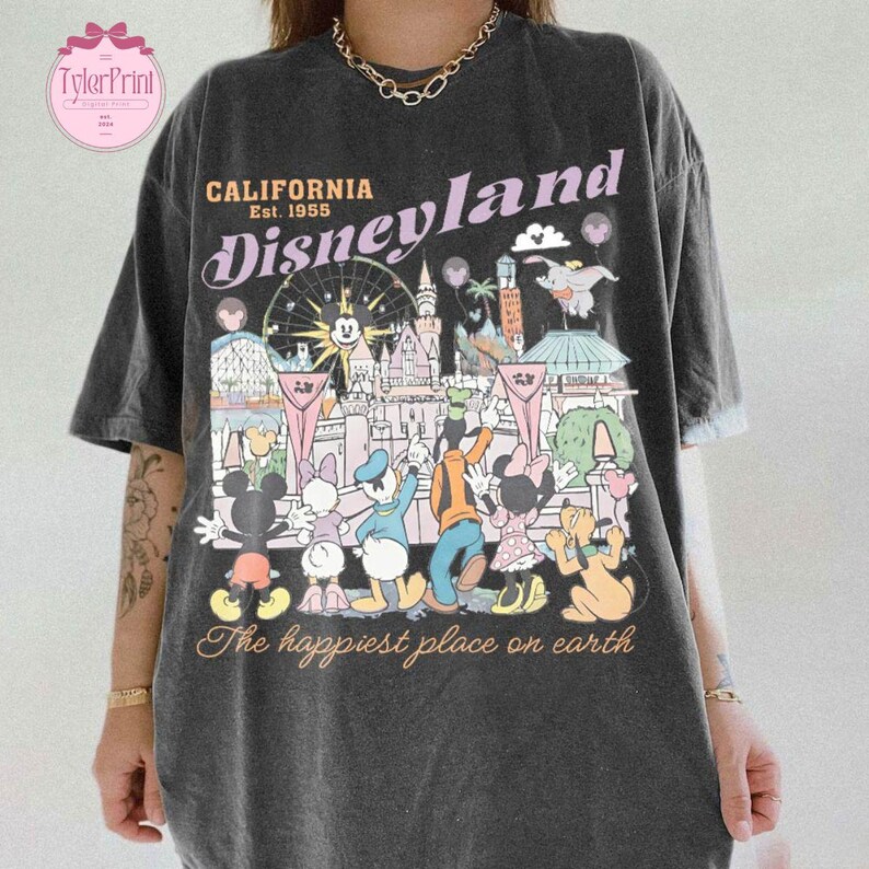 Puede incluir: Camiseta gris oscuro con un gr&aacute;fico colorido de Disneyland. El dise&ntilde;o incluye el texto "California Disneyland Est. 1955" y una representaci&oacute;n de varios personajes y lugares emblem&aacute;ticos de Disney. Tambi&eacute;n se incluye la frase "The happiest place on earth".