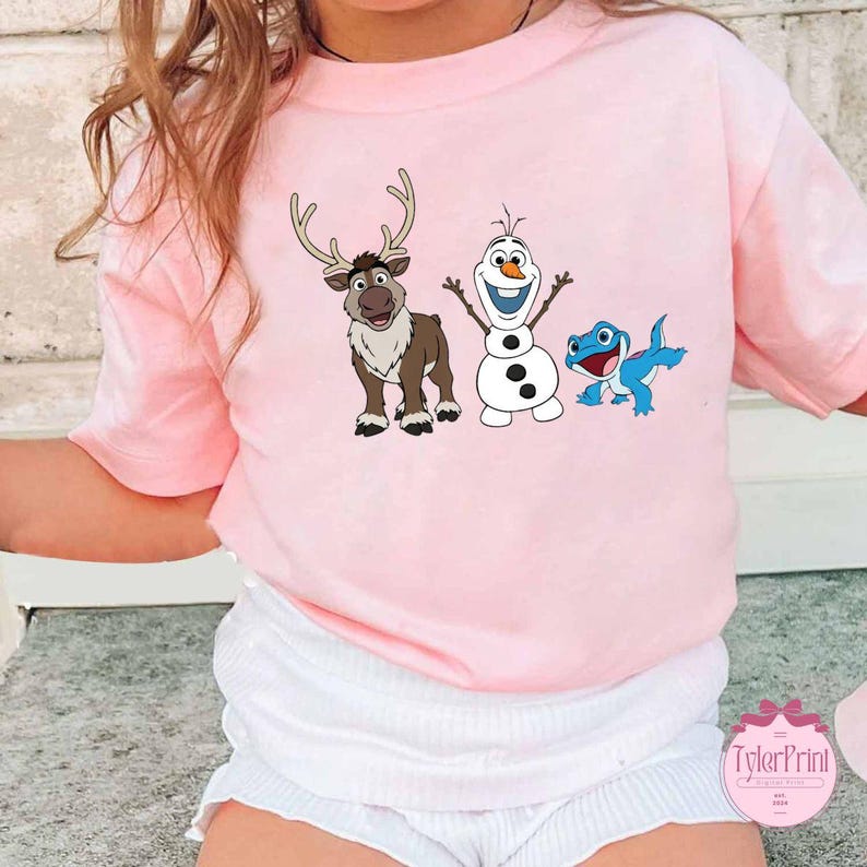 Peut inclure: Un t-shirt rose clair avec des personnages de dessins anim&eacute;s du film La Reine des Neiges. Le motif comprend un renne, un bonhomme de neige et un l&eacute;zard bleu. Le t-shirt est &agrave; manches courtes.