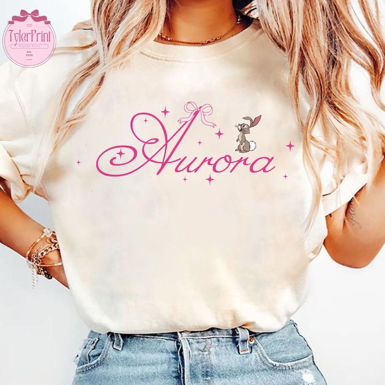 Pu&ograve; includere: Una maglietta bianca con un font corsivo rosa che dice "Aurora" con un fiocco rosa e un piccolo coniglio dei cartoni animati sopra la parola.