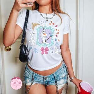 May include: White t-shirt featuring a cartoon image of Olaf from Frozen, holding a blue bird. The design includes snowflakes, leaves, and a pink bow. The shirt has a crew neck and short sleeves.