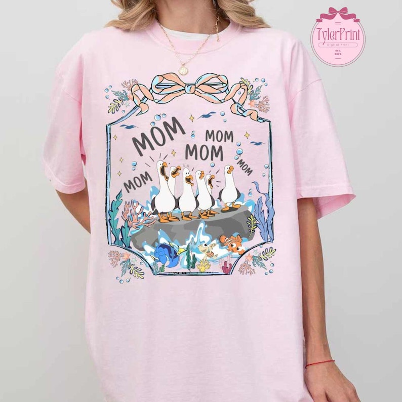 May include: A light pink t-shirt featuring a cartoon design with the word "MOM" repeated around a group of geese. The geese are standing on a rock in an underwater scene with fish and seaweed. A bow and floral elements frame the design.