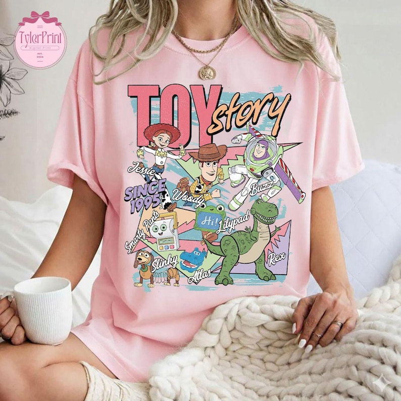 K&ouml;nnte beinhalten: Ein hellrosa T-Shirt mit einem farbenfrohen Grafikdruck von Toy Story-Figuren wie Woody, Buzz Lightyear, Jessie, Rex und Slinky. Der Text lautet TOY story SINCE 1995, Smarty Pants, Lilypad und Atlas in verschiedenen Schriftarten.