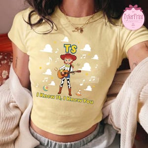 May include: A pale yellow short-sleeved t-shirt featuring a graphic of a red-haired cowgirl playing an acoustic guitar. The design includes the text TS and I Knew It, I Knew You, surrounded by white clouds, musical notes, a ball, and a traffic cone.