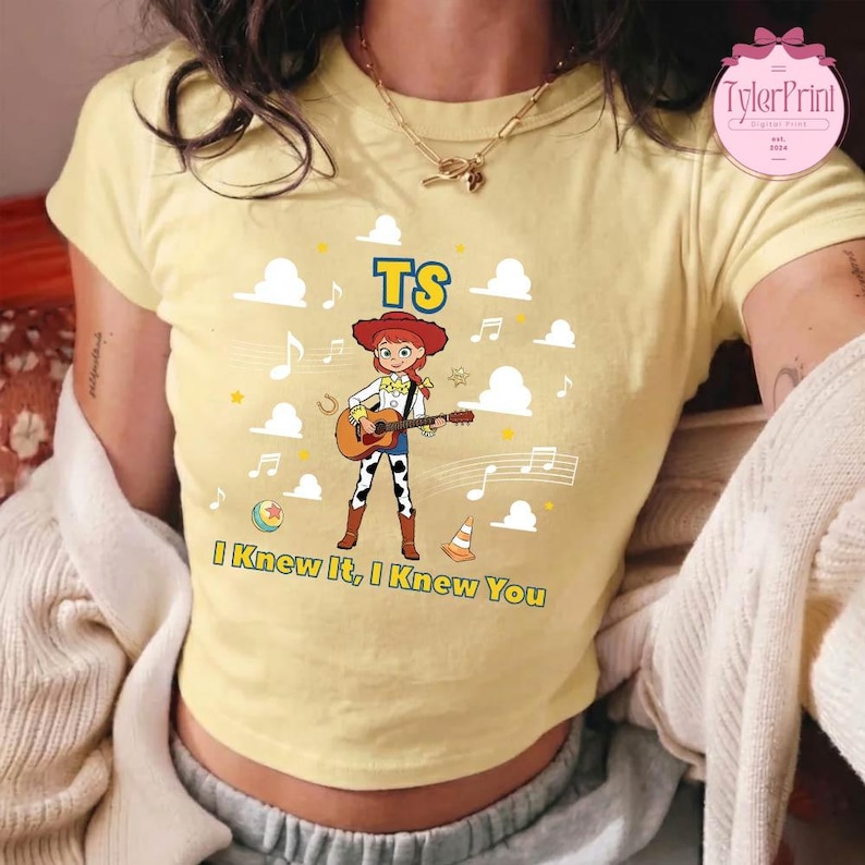 May include: A pale yellow short-sleeved t-shirt featuring a graphic of a red-haired cowgirl playing an acoustic guitar. The design includes the text TS and I Knew It, I Knew You, surrounded by white clouds, musical notes, a ball, and a traffic cone.