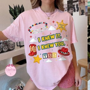 May include: A light pink t-shirt featuring a graphic print with a colourful beaded bracelet, a yellow sheriff star badge with the text Infinity and Beyond, red cowboy boots, a red cowboy hat, and blocks spelling TS5. The text reads I KNEW IT, I KNEW YOU.