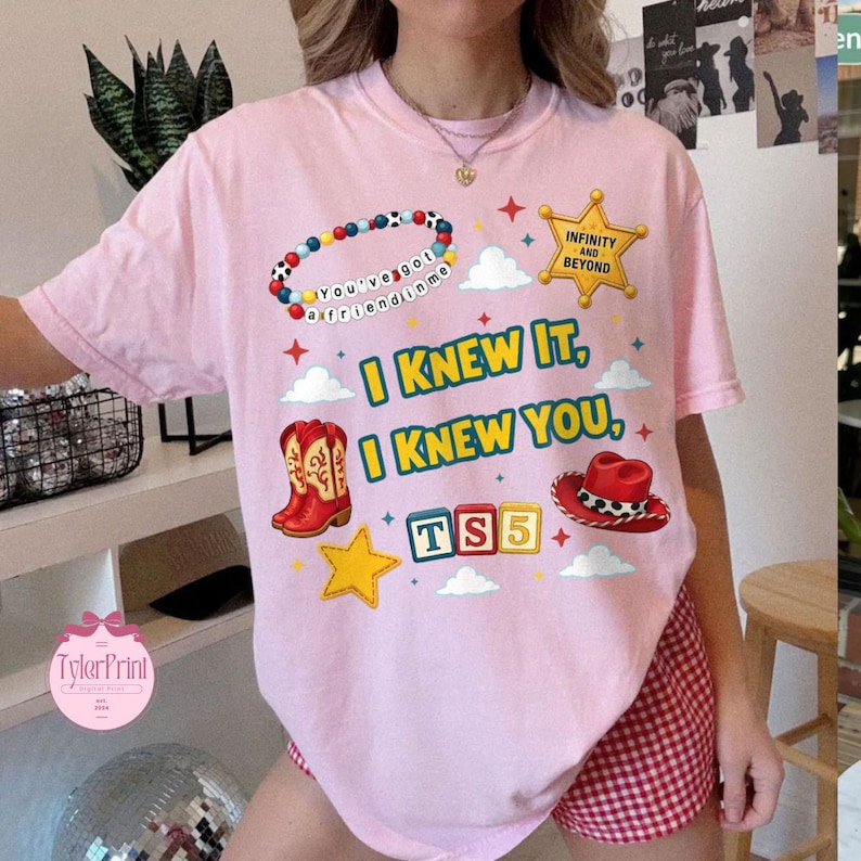 May include: A light pink t-shirt featuring a graphic print with a colourful beaded bracelet, a yellow sheriff star badge with the text Infinity and Beyond, red cowboy boots, a red cowboy hat, and blocks spelling TS5. The text reads I KNEW IT, I KNEW YOU.