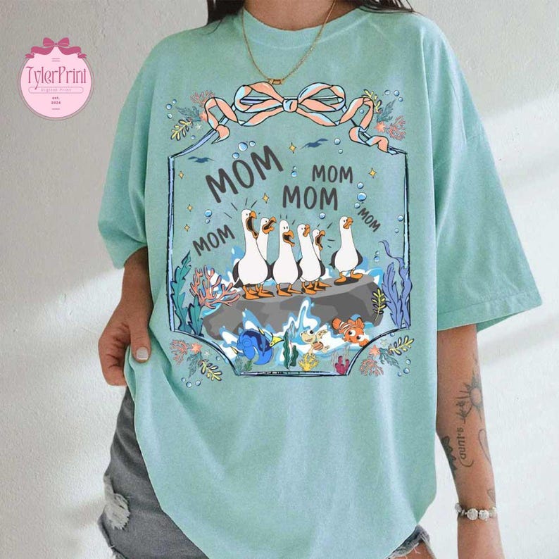May include: A light teal t-shirt with a graphic featuring five white geese on a rock, surrounded by aquatic plants and the word "MOM" repeated. A pink bow and the words "TylerPrint" are in the upper left corner.