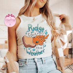May include: A cream-coloured vest top featuring a cartoon graphic of an orange fish with blue spots and the text "Pudge Controls the Weather" in blue. The fish is holding a piece of toast. The top is worn by a person.