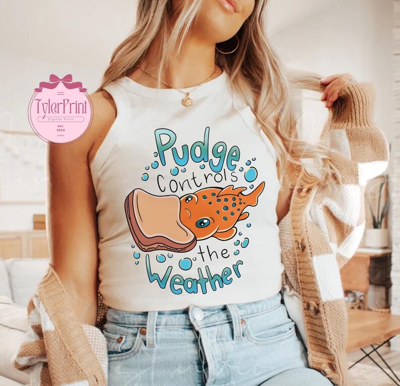 May include: A cream-coloured vest top featuring a cartoon graphic of an orange fish with blue spots and the text "Pudge Controls the Weather" in blue. The fish is holding a piece of toast. The top is worn by a person.