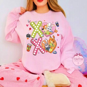 May include: Pink sweatshirt featuring the characters from Zootopia. The design includes two "XO" symbols, each with a heart and character illustrations. The sweatshirt also has coffee cups and other small graphics.