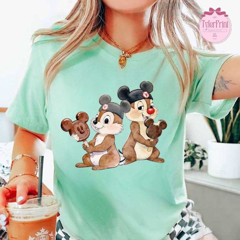 Puede incluir: Camiseta verde claro con una ilustraci&oacute;n de dibujos animados de dos chipmunks sosteniendo helados con forma de Mickey Mouse. Los chipmunks llevan sombreros negros con detalles rojos. La camiseta tiene el texto "TylerPrint" en un c&iacute;rculo rosa.