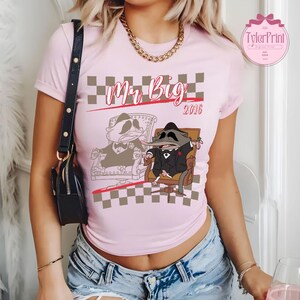 May include: A pale pink t-shirt with a cartoon raccoon in a suit, seated in a chair. The text "Mr. Big 2016" is above the raccoon. The shirt has a checkered pattern and is being worn by a person.