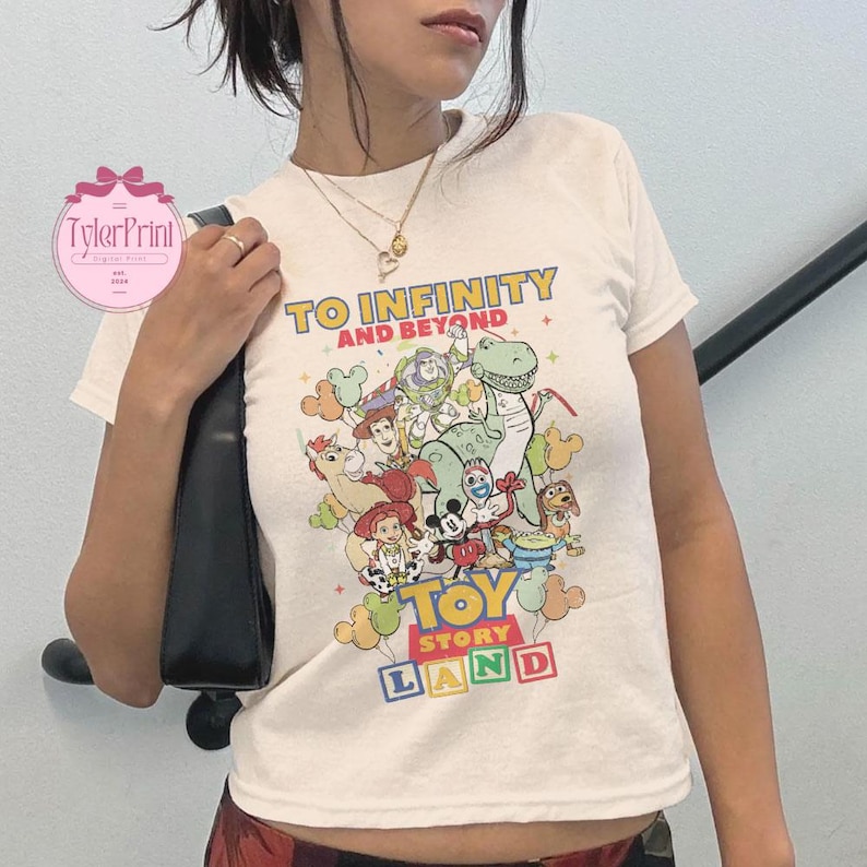 以下が含まれることがあります： クリーム色のTシャツ。カラフルなトイ・ストーリーのキャラクターのグラフィックが描かれています。「TO INFINITY AND BEYOND」と「TOY STORY LAND」の文字が入っています。半袖のシャツです。