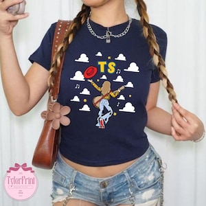 May include: A navy blue short-sleeved t-shirt featuring a graphic of a person with long hair wearing a yellow plaid shirt and cow-print bell-bottoms, holding a red hat and guitar, surrounded by white clouds, musical notes, and the yellow text TS.