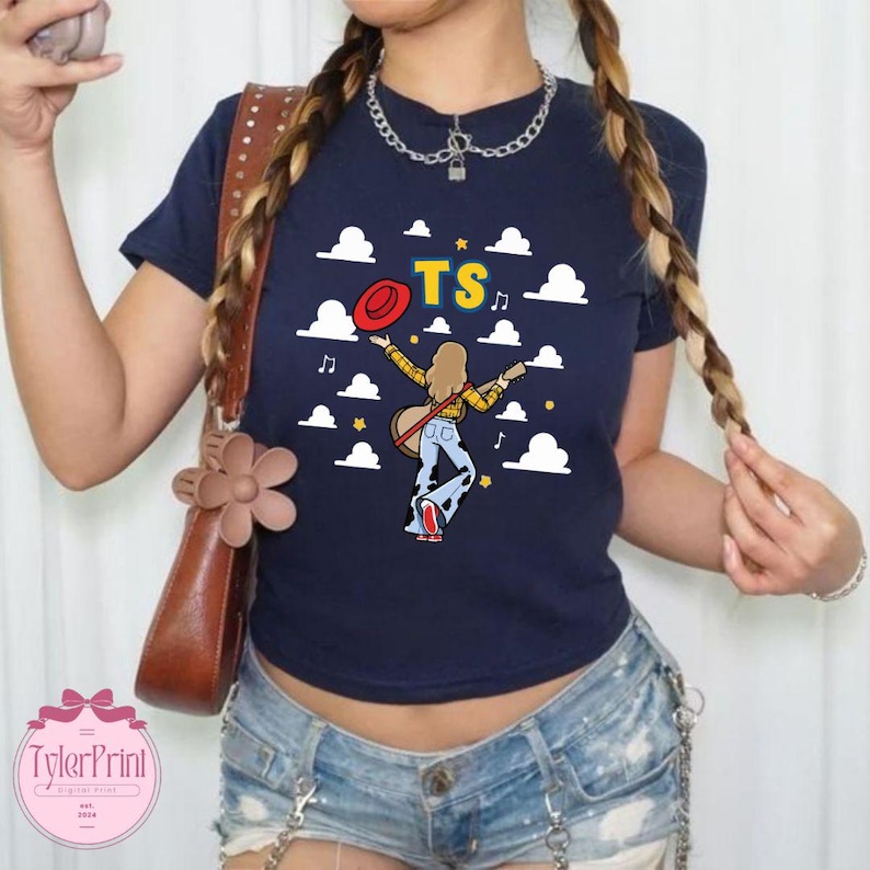 May include: A navy blue short-sleeved t-shirt featuring a graphic of a person with long hair wearing a yellow plaid shirt and cow-print bell-bottoms, holding a red hat and guitar, surrounded by white clouds, musical notes, and the yellow text TS.