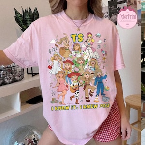 May include: A light pink t-shirt featuring a colorful graphic print of various animated characters in a cartoon style. The text TS is printed at the top, and the text i knew it, i knew you is printed at the bottom. The design includes musical notes, stars, and small toys.