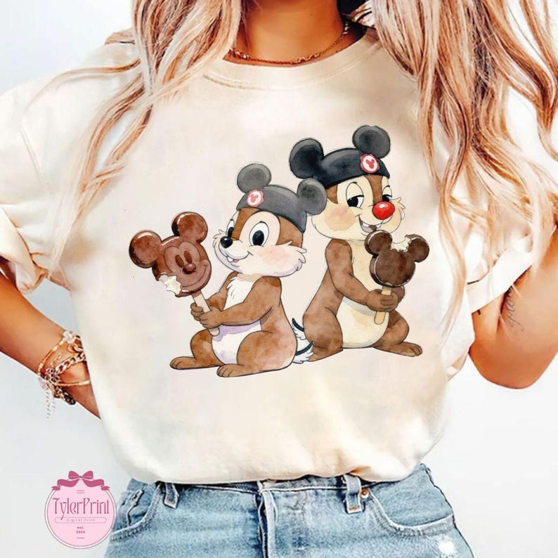 Puede incluir: Camiseta color crema con una ilustraci&oacute;n en acuarela de dos ardillas de dibujos animados, cada una sosteniendo un helado con forma de Mickey Mouse. Las ardillas llevan sombreros negros con un coraz&oacute;n rojo. La camiseta tiene un aspecto vintage.
