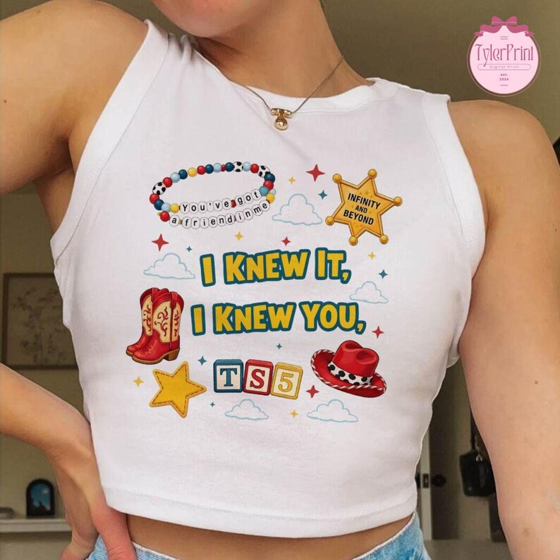 May include: A white sleeveless tank top featuring a graphic print with the text I KNEW IT, I KNEW YOU, TS5, INFINITY AND BEYOND, and YOU'VE GOT A FRIEND IN ME. The design includes a beaded bracelet, a sheriff star, red cowboy boots, a hat, and stars.