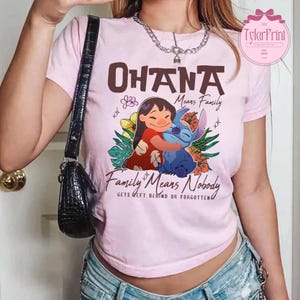 May include: A light pink t-shirt featuring a graphic of Lilo and Stitch hugging, with the text "Ohana Means Family" and "Family Means Nobody Gets Left Behind or Forgotten". The design includes flowers and tropical leaves.