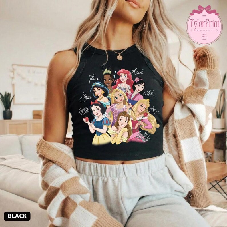 May include: Black tank top featuring a colorful graphic of Disney princesses, including Snow White, Ariel, and Belle. The princesses' names are written above them in a cursive font. The word "BLACK" is printed at the bottom.