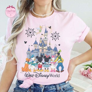 Puede incluir: Camiseta rosa claro con un gr&aacute;fico colorido del castillo de Walt Disney World, fuegos artificiales y personajes de Disney. El texto "Walt Disney World" est&aacute; impreso en una fuente cursiva en la parte inferior.