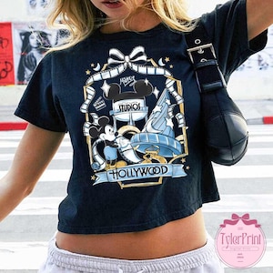 May include: Black t-shirt featuring a colorful graphic of Mickey Mouse, the Hollywood sign, and the words "Mickey Studios Hollywood." The design includes film reels, stars, and a bow. The shirt has short sleeves.