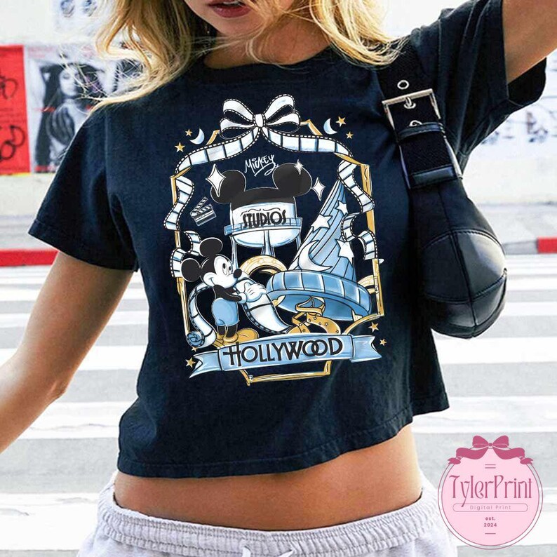 May include: Black t-shirt featuring a colorful graphic of Mickey Mouse, the Hollywood sign, and the words "Mickey Studios Hollywood." The design includes film reels, stars, and a bow. The shirt has short sleeves.