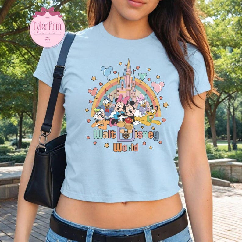 May include: A light blue cropped t-shirt with a graphic of Mickey Mouse and friends in front of the Cinderella Castle at Walt Disney World. The graphic is surrounded by hearts, stars, and balloons. The text "Walt Disney World" is printed below the graphic.