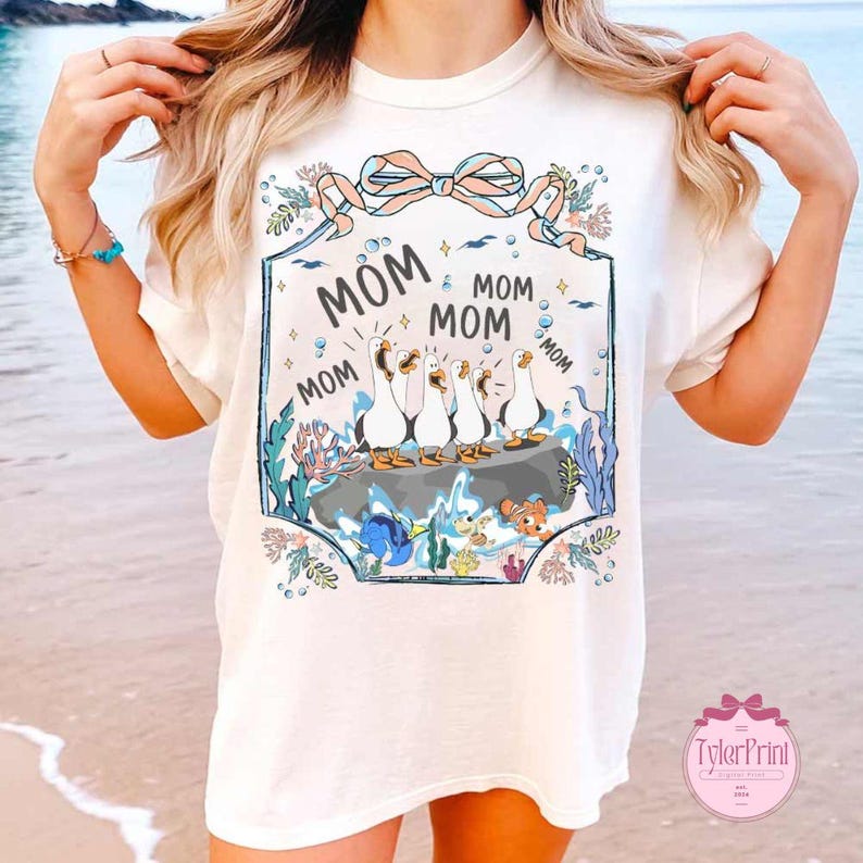 May include: Cream-colored t-shirt featuring a whimsical design with the word "MOM" repeated around a group of cartoon ducks. The design includes aquatic elements like coral, fish, and bubbles, with a decorative bow at the top.